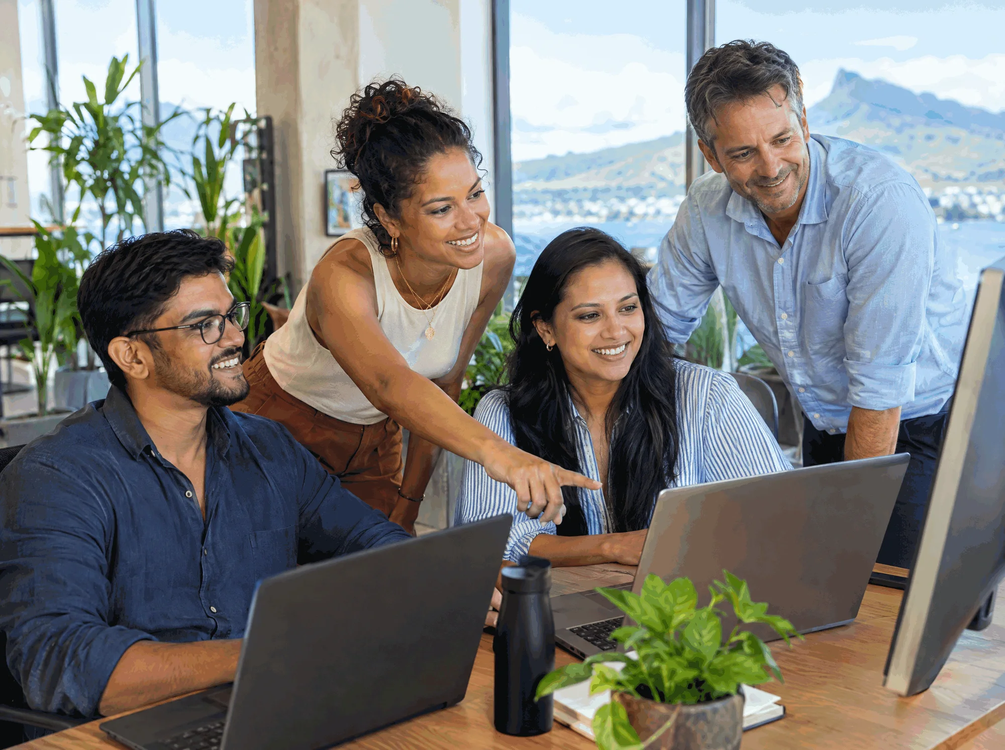 Four colleagues smiling at a laptop in a bright office, one pointing at the screen, mountains and the sea visible through the windows behind them.